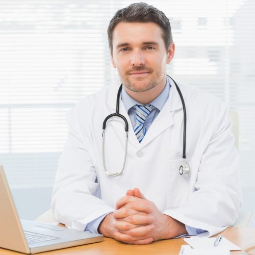 Portrait of a smiling male doctor with laptop sitting at desk in medical office